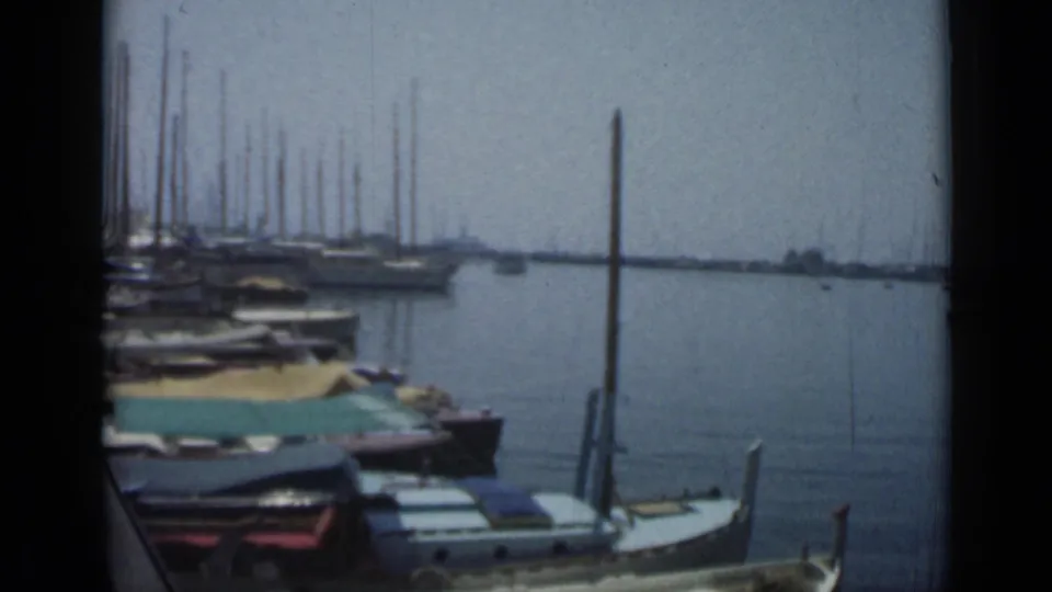 1962 Cannes harbor footage with covered boats near the dock, sailboat masts to the left, open water, and soft 8mm color texture.