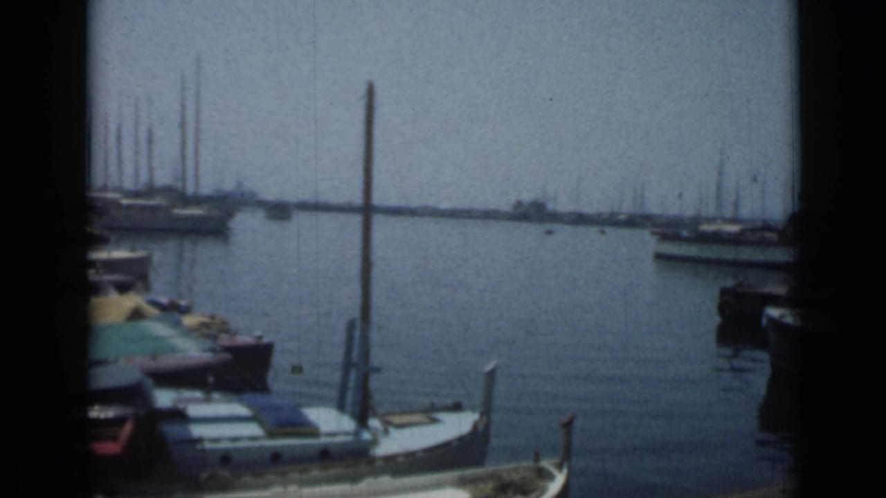 1962 Cannes harbor view with small moored boats, calm water, distant sailboat masts, and a hazy waterfront horizon.
