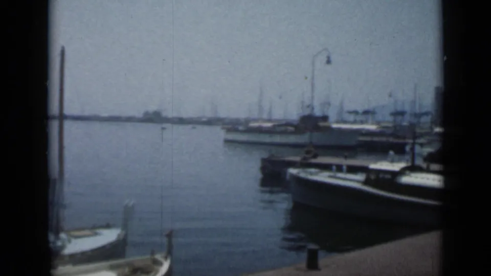 1962 Cannes marina view with small sailboats, motorboats, a dock edge, curved lamps, and rows of distant masts under a hazy sky.