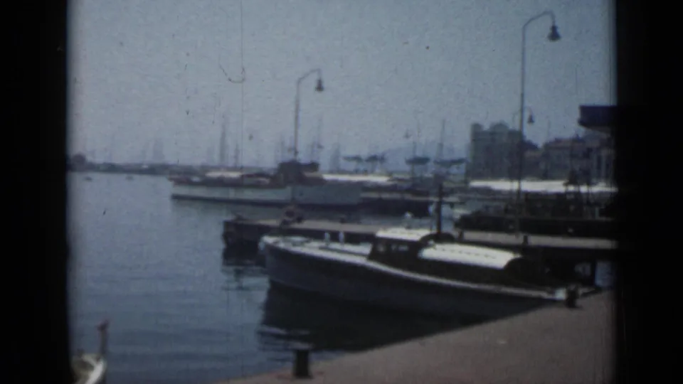 Cannes harbor scene from 1962 with docked boats beside the quay, curved lamps, distant masts, and low waterfront buildings.