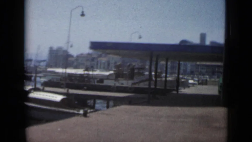 1962 Cannes harbor view with a covered quay, moored boats, calm water, and distant waterfront buildings on 8mm film.