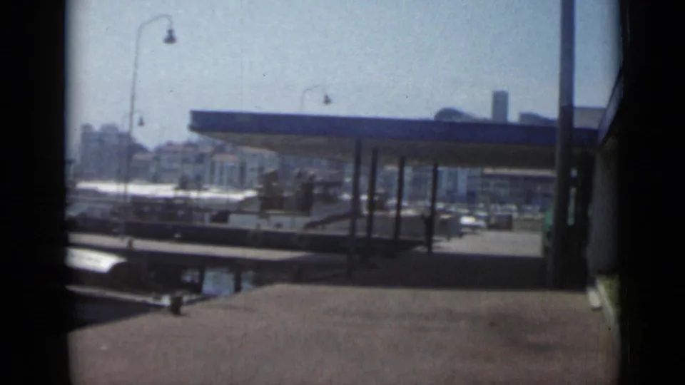 1962 Cannes harbor view with a dockside shelter, small boats, street lamps, and waterfront buildings in soft 8mm color.