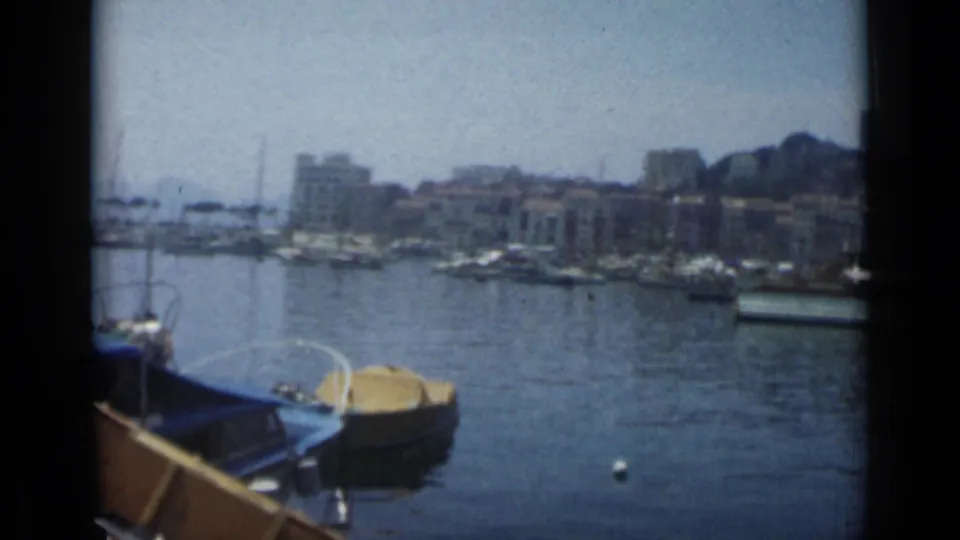 Wide 1962 Cannes marina view with calm water, a yellow-covered boat, moored vessels, and waterfront buildings beyond.