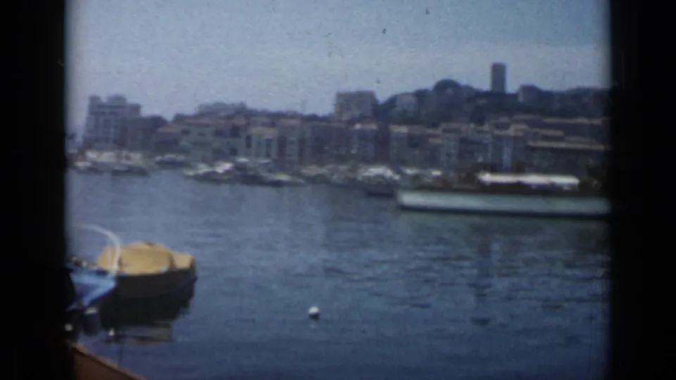 1962 Cannes harbor view with a yellow-covered boat in the foreground, boats along the quay, and hillside buildings behind.