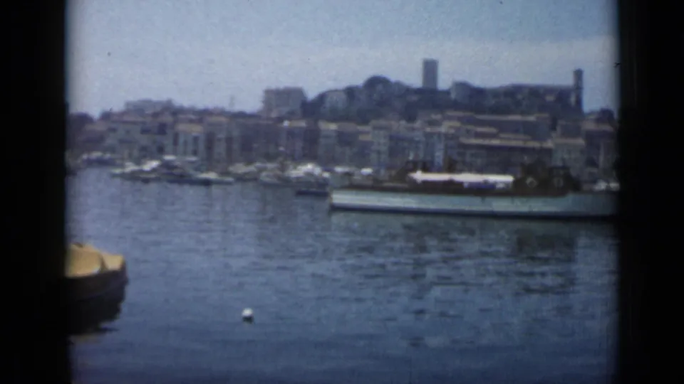1962 8mm view across Cannes harbor toward moored boats, dense waterfront buildings, and a tower on the hillside.