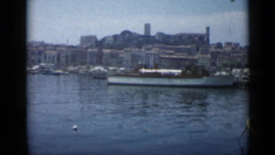 1962 Cannes harbor scene with small white boats, a larger vessel near shore, and hillside buildings in soft 8mm color.