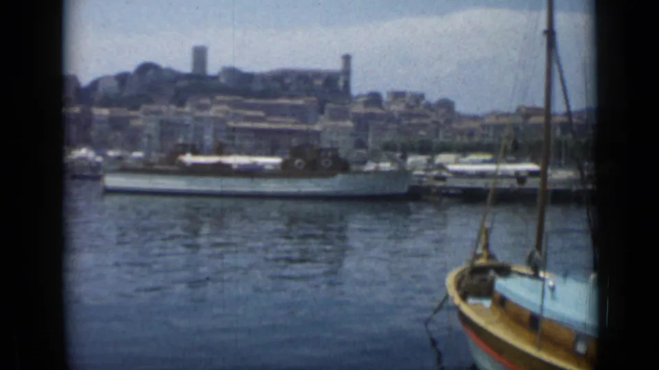 1962 8mm Cannes harbor frame showing boats on calm water, city buildings on the shore, and a nearby small boat at the right edge.