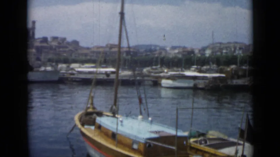 Vintage 8mm view of Cannes harbor with a wooden sailboat, marina boats, dark water, and softened hillside buildings.