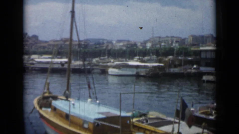 1962 Cannes marina frame with a foreground sailboat, moored boats, waterfront buildings, and visible 8mm grain.