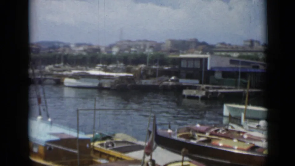 Cannes marina footage shows small boats tied along the waterfront, with low buildings and hillside structures in the distance.