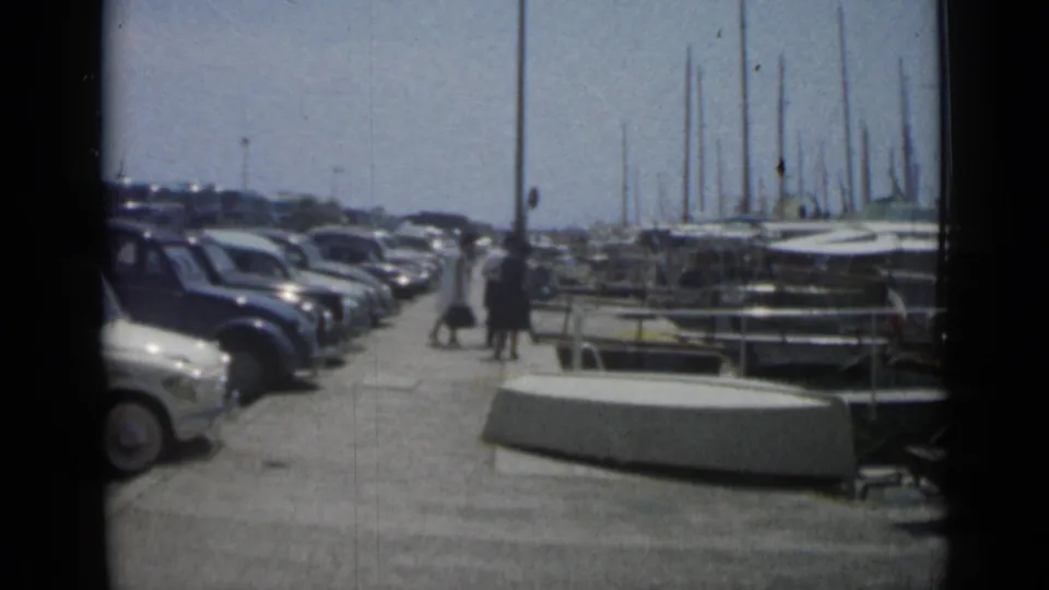 At the Cannes marina, a row of vintage cars stands beside docks and sailboat masts while two distant pedestrians walk along the waterfront.