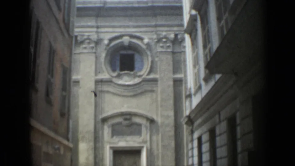 1962 Cannes home-movie frame looking up a narrow alley toward an ornate stone facade with a circular upper window.