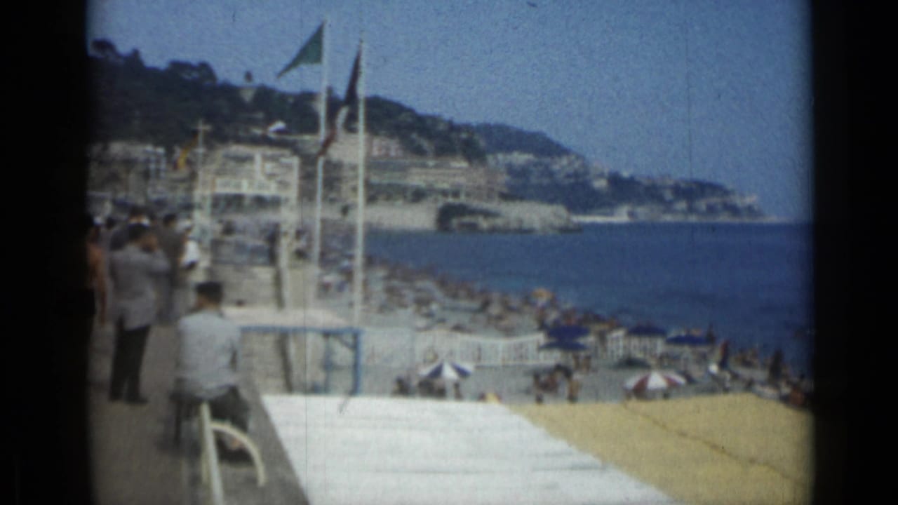 Cannes beach with sunbathers, umbrellas, flags, sea, and hillside buildings.