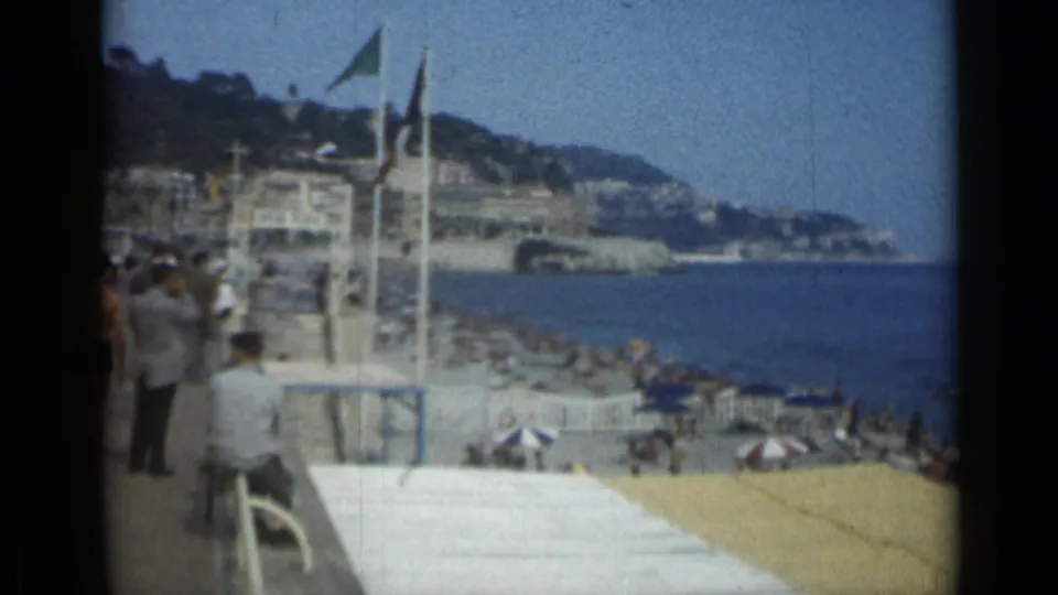 1962 Cannes beach view with flags, sunbathers, striped umbrellas, blue sea, and hillside waterfront buildings.
