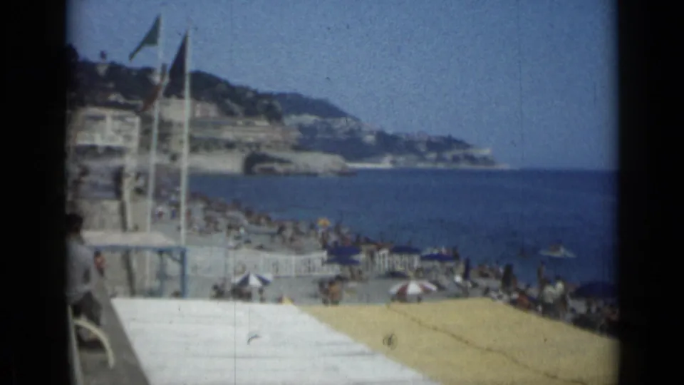 1962 Cannes shoreline with beachgoers under umbrellas, flags near the promenade, swimmers, and the Mediterranean beyond.