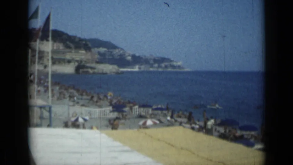 1962 Cannes beach and sea seen from above, with sunbathers, striped umbrellas, swimmers, and coastal buildings in the distance.