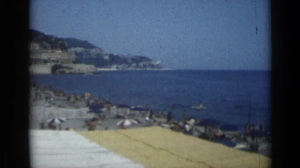 1962 Cannes beach scene with sunbathers, striped umbrellas, small swimmers in the blue sea, and hillside coastline beyond.