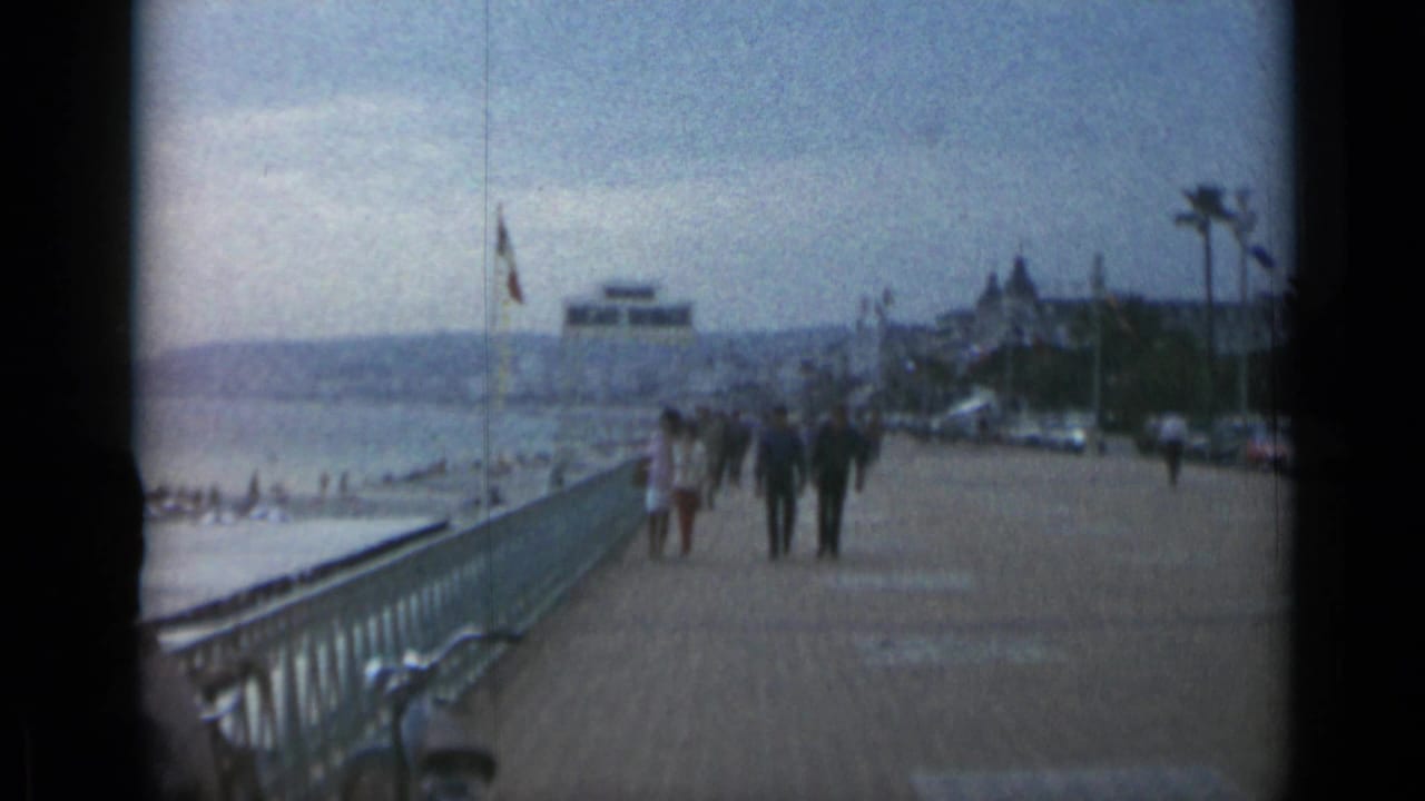 Pedestrians walk along a seaside promenade with railings, flags, beach, cars, and distant buildings.