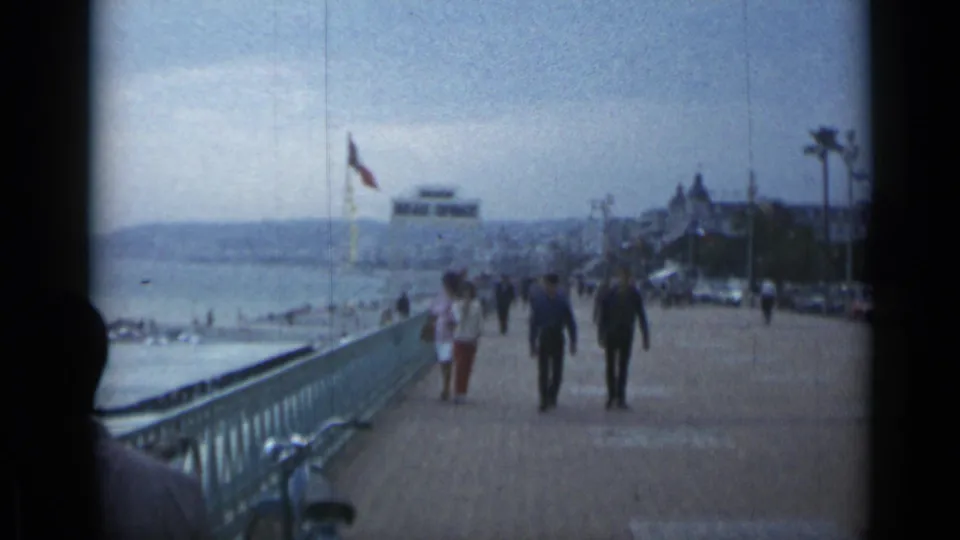 Cannes waterfront in 1962 with two men prominent on the promenade, other walkers, beach and water at left, flags, cars, and palm trees.