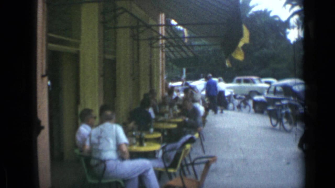 Outdoor cafe tables with seated patrons, parked cars, bicycles, and awnings along a sidewalk.