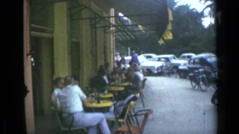 Cannes sidewalk cafe view from 1962 with patrons seated under awnings, bicycles near the curb, and classic cars parked beyond.