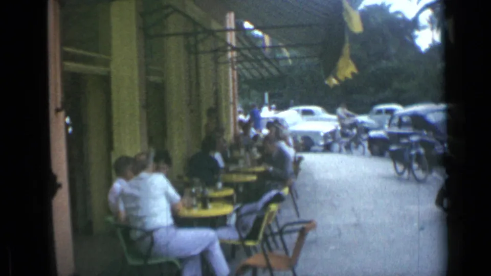 1962 Cannes cafe patrons sit at yellow sidewalk tables while classic cars and bicycles line the street beyond the awning.