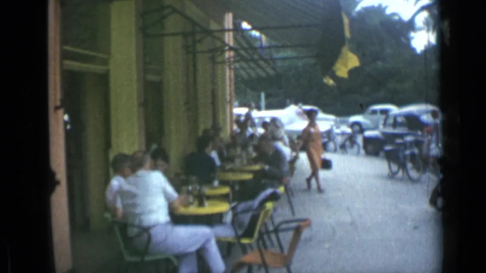 In the supplied 1962 Cannes footage, a woman in a light dress walks past sidewalk cafe patrons, bicycles, and parked classic cars.