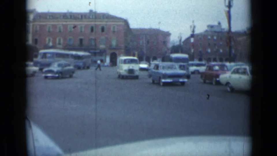 Cannes, France 1962 traffic flow with buses, compact cars, pedestrians, streetlamps, and a brick building backdrop.
