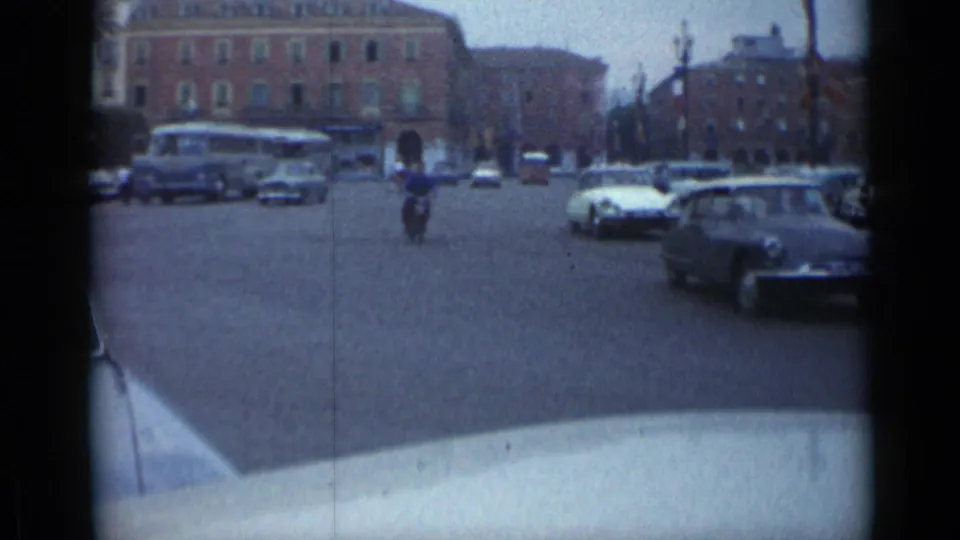 1962 Cannes street traffic with a bus, several cars, pedestrians, and a motorcycle or scooter rider in a broad intersection.