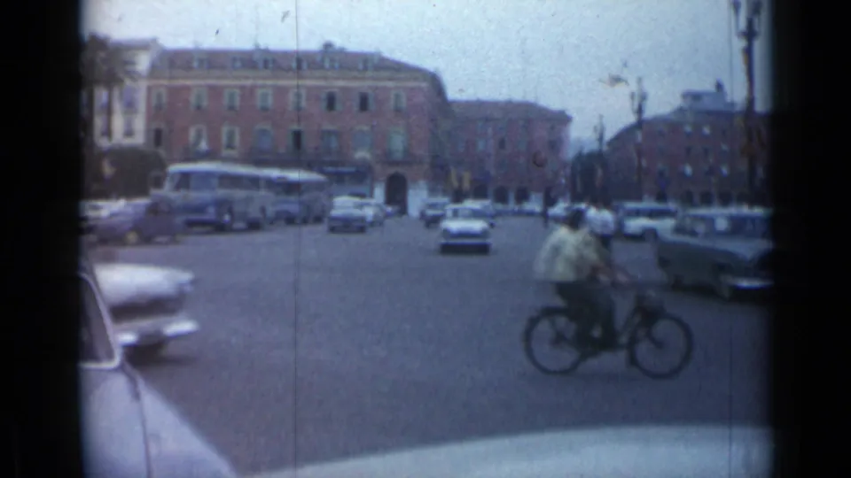 A cyclist rides across the 1962 Cannes intersection with period cars, a bus, and multi-story buildings in the background.
