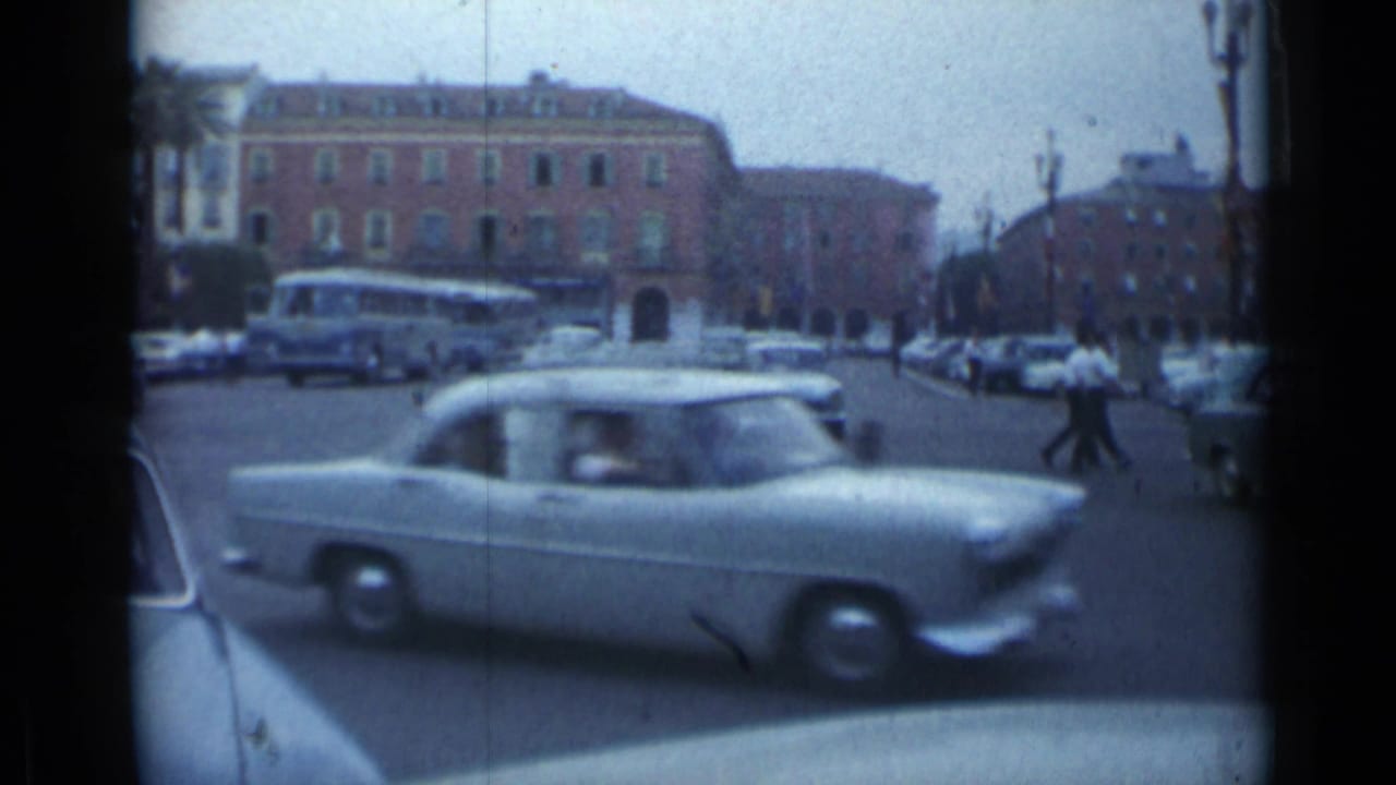 Classic car passing across a busy city avenue with bus and pedestrians in the background.
