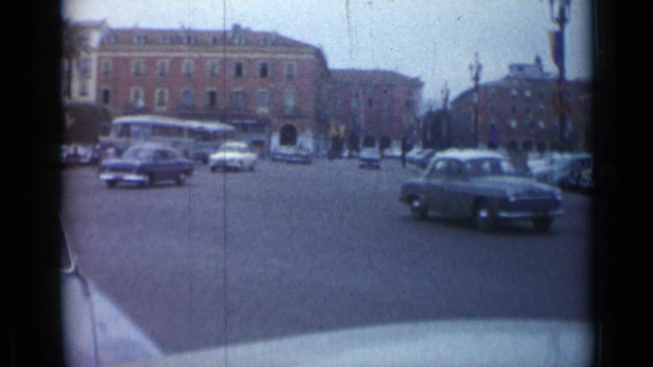 Cars moving through a city square with mid-century buildings in the background.