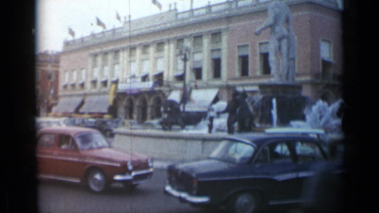 Cars pass a fountain and statue in front of a columned public building.