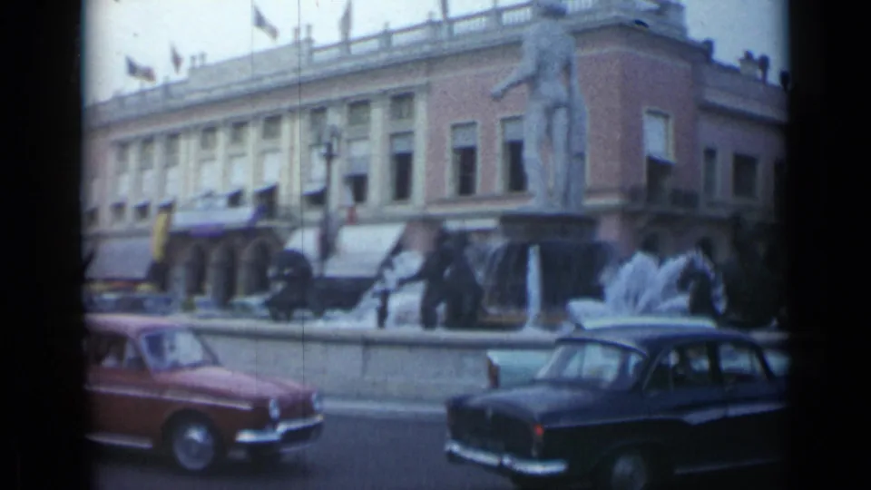 1962 Cannes traffic scene with a red car, dark sedan, fountain spray, statues, and pedestrians near a civic building.
