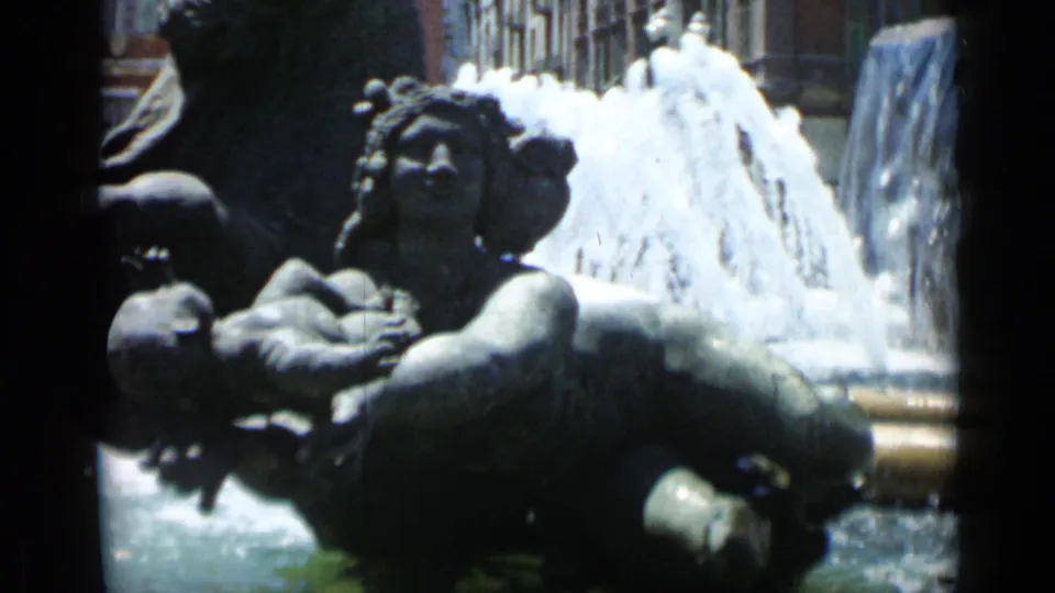 1962 Cannes 8mm frame showing bronze fountain figures, including a woman holding a child-shaped sculpture, with water behind.
