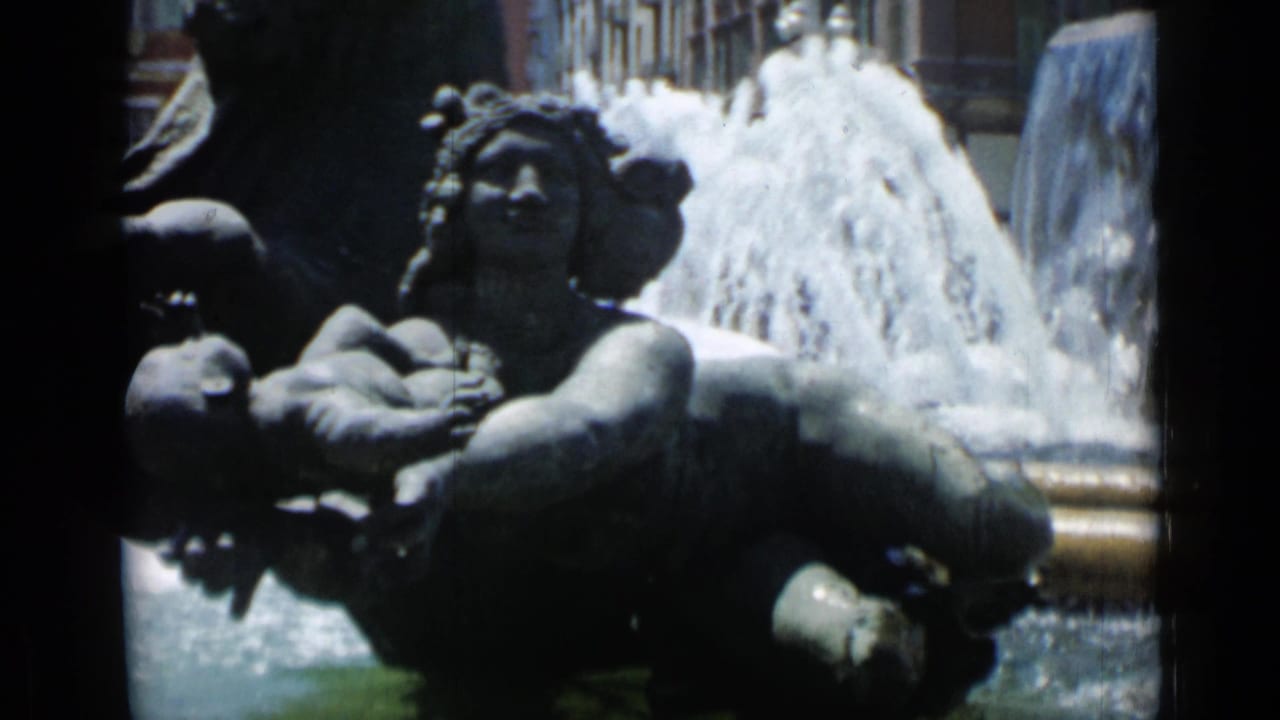 Stone fountain sculpture with human figures and water spray behind it.