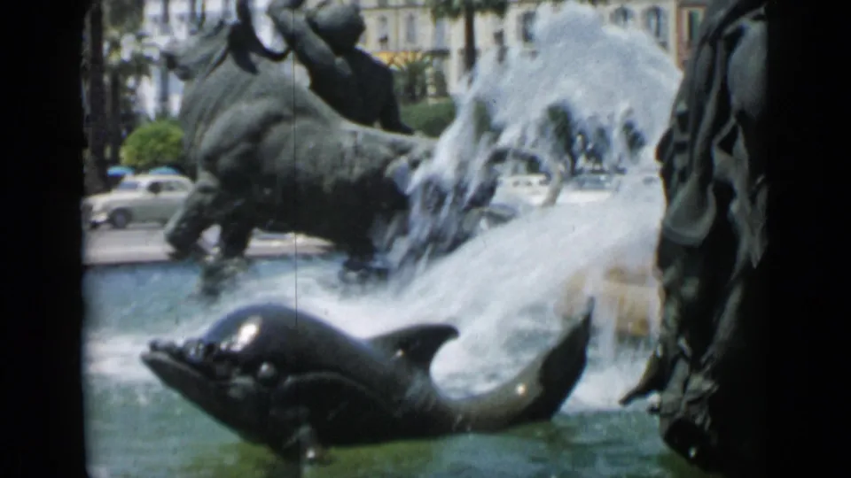 Cannes 1962 8mm frame showing a dolphin fountain sculpture, water jets, cars, palm trees, and pale buildings behind.