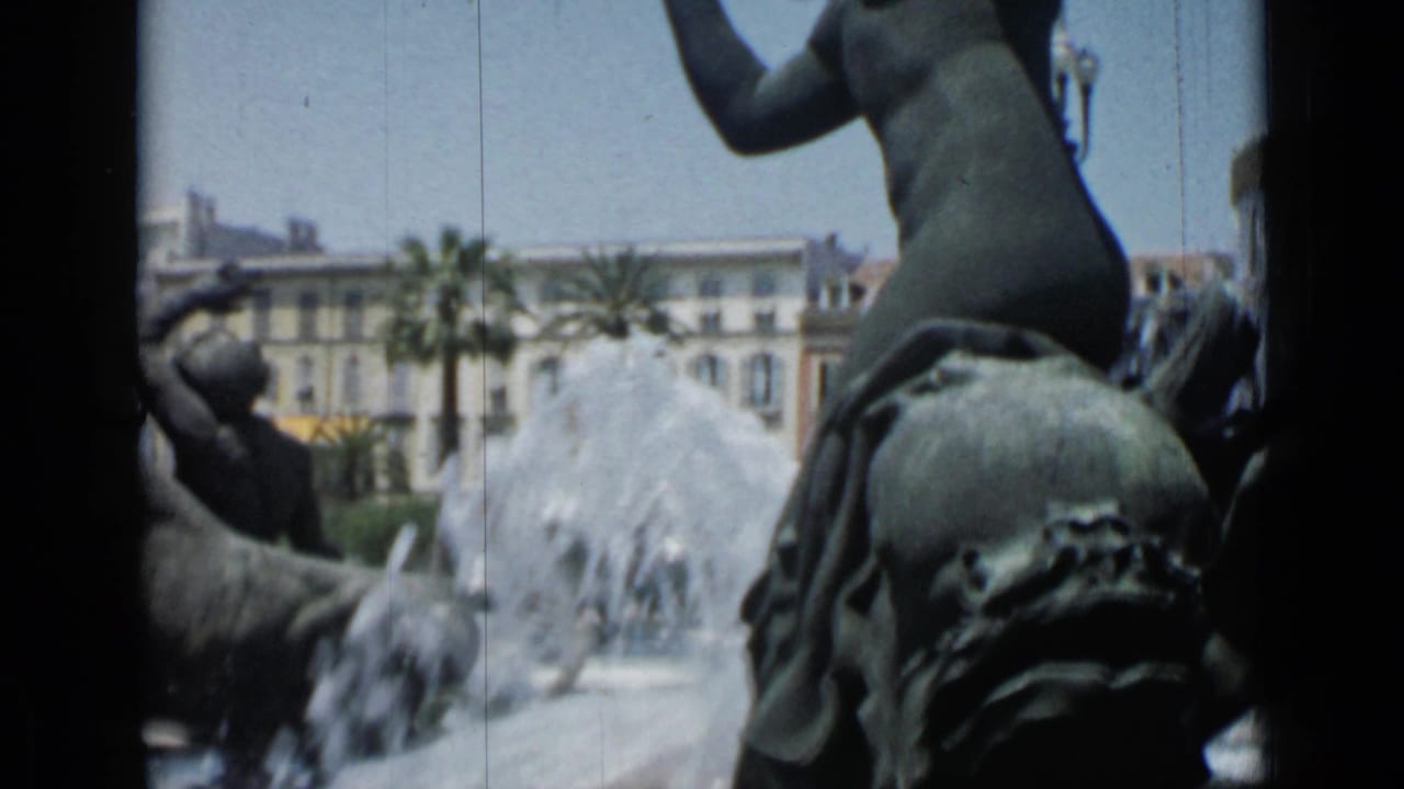Bronze fountain sculpture with water spray, palm trees, and buildings behind it.