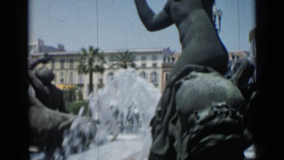 1962 Cannes home-movie frame showing a bronze fountain sculpture with water jets, palm trees, and sunlit buildings in the background.
