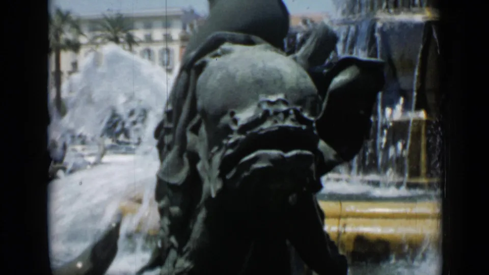 1962 Cannes 8mm frame showing the fountain's bronze horse form close up, with spray, basin detail, and pale buildings behind.