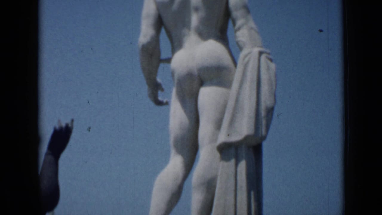 Rear close view of a nude stone male statue against blue sky with a partial hand at the edge.