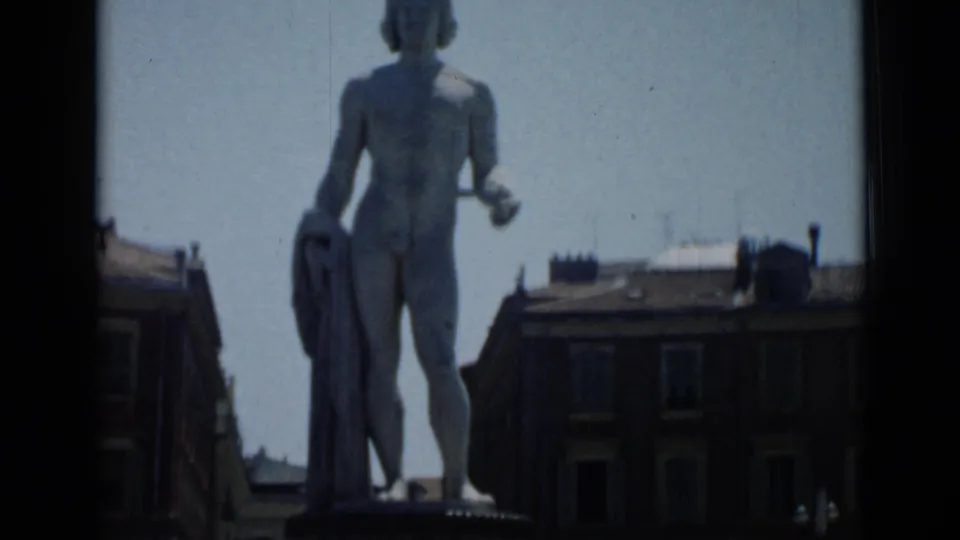 Front view of the nude marble statue in Cannes with surrounding building roofs visible in the 1962 8mm frame.