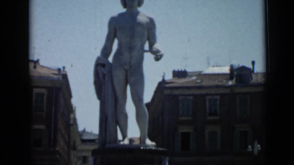 The Cannes fountain statue appears centered between urban facades, with soft 8mm color and slight handheld framing.