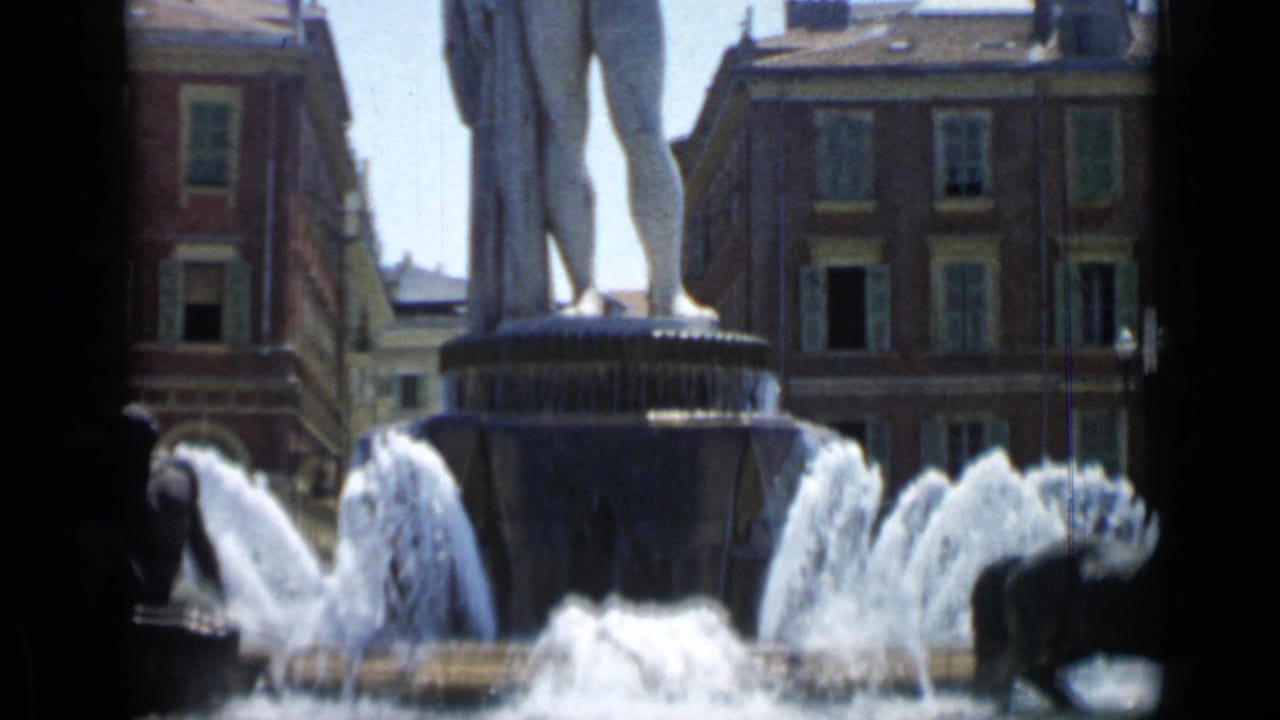 Fountain with spraying water, sculpture base, and tall statue legs before city buildings.