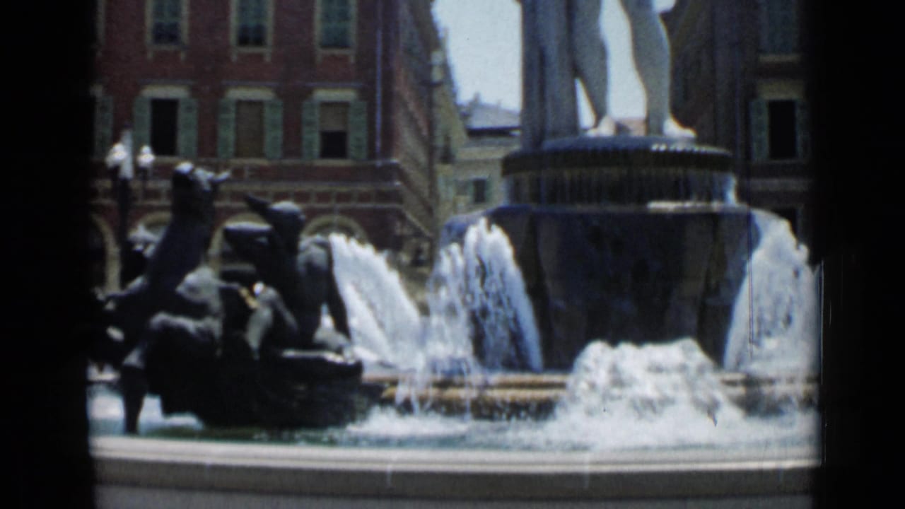 1962 Cannes frame of a fountain sculpture with horse forms, splashing water, and shuttered urban facades behind.