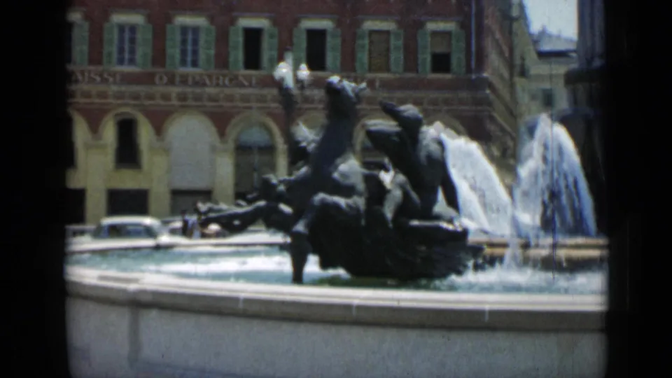 1962 Cannes 8mm view of a horse fountain sculpture, spraying water, storefront arches, and a vintage car in the background.