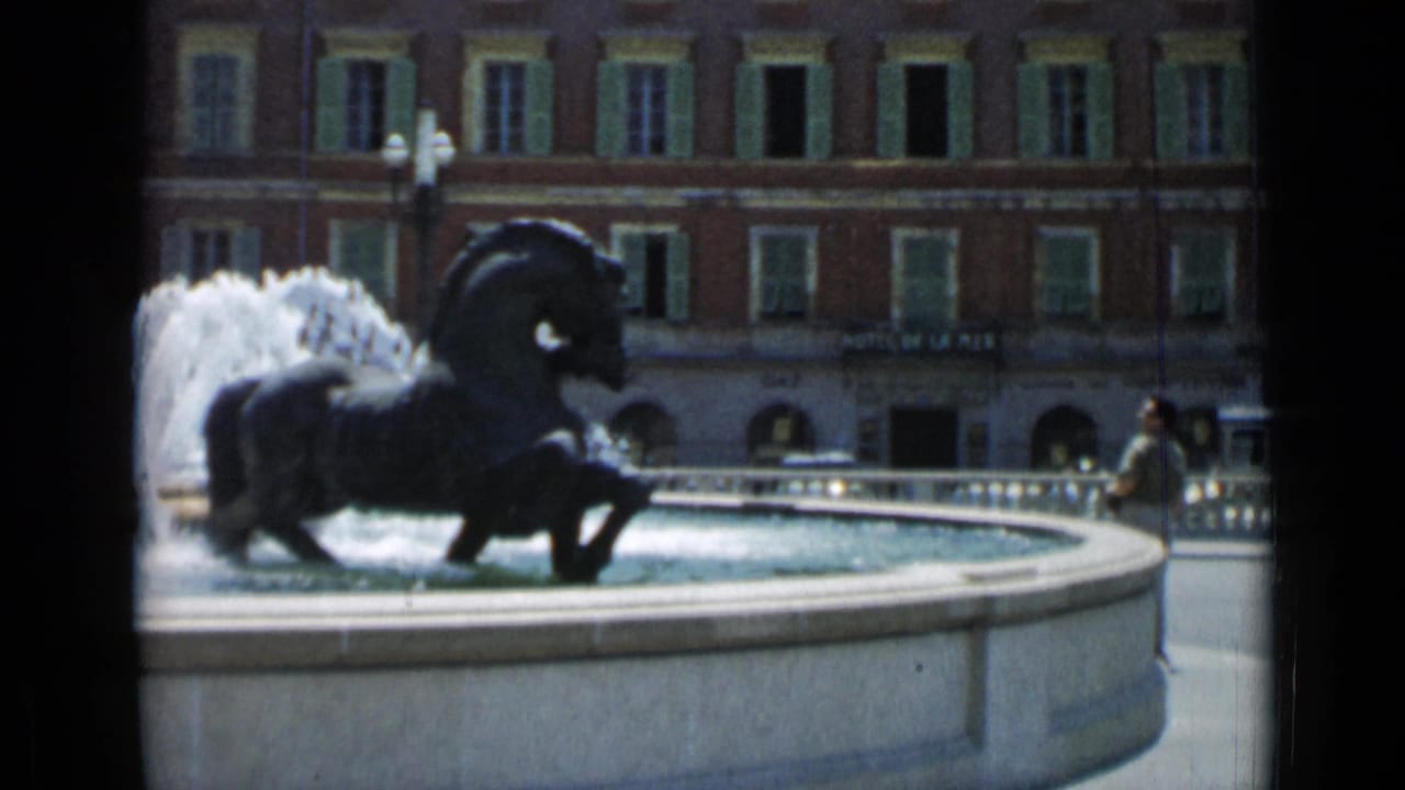 Horse fountain with water spray in a town square, with buildings and pedestrians behind it.