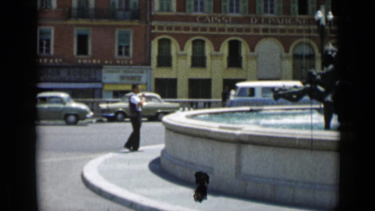 Man standing near a fountain while cars and a van pass in front of a bank building.