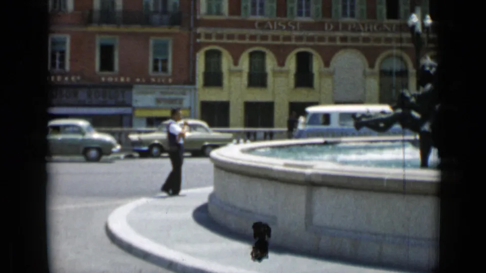 Man in a white shirt stands near a Cannes fountain as 1960s cars and a light van pass by the CAISSE D'EPARGNE building.