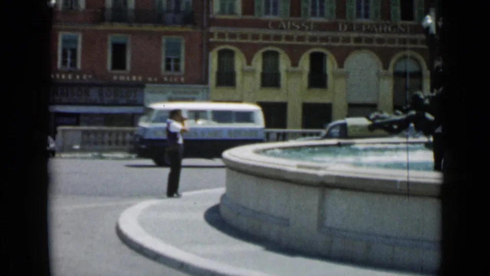 1962 Cannes frame showing a man near the fountain edge, with a passing van, classic cars, and storefront signage across the square.
